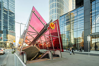 Entrance to the metro station with red glass roof next to modern residential and commercial buildings in Warsaw.